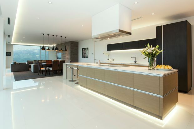 A bright, spacious open-plan kitchen and dining/living area. The kitchen features a large central island with pale timber-look cabinetry, a light stone benchtop, integrated appliances, a stainless-steel tap and sink, and a prominent white rangehood canopy. Glossy light flooring, white walls and ceiling, dark horizontal joinery, and concealed LED lighting create a crisp contemporary interior. A dining table, pendant lights and living area sit beyond the kitchen, with large glazing at the rear.