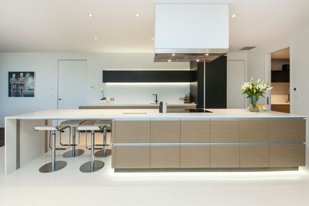 A bright, contemporary open-plan kitchen dominated by a large island with integrated seating, pale timber-look cabinetry, a white benchtop, and a black feature wall of cabinetry. Recessed lighting and under-cabinet illumination create a clean, streamlined appearance. A dining or living area is not clearly visible.