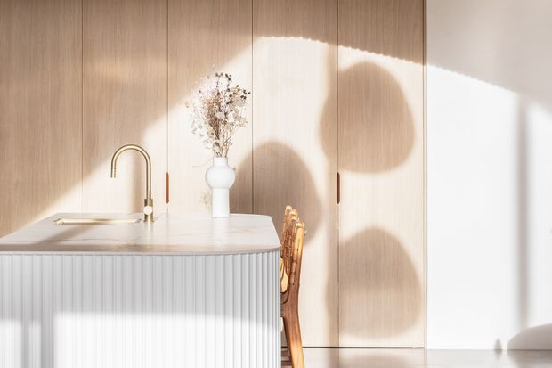 Bright minimalist kitchen interior with a pale stone island featuring a vertically fluted front, light timber full-height cabinetry, and a slim brass gooseneck tap. A woven timber chair and ceramic vase with dried branches add natural warmth, while strong sunlight creates graphic shadows across the cabinetry and floor.