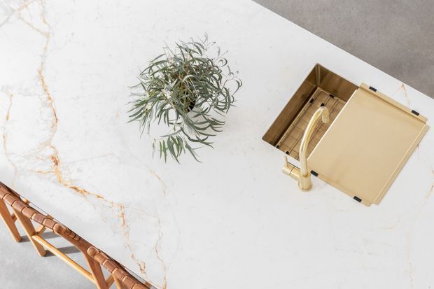 Top-down view of a bright marble kitchen island or benchtop with subtle grey veining and warm bronze-gold accents. A leafy plant, open tan notebook or folder, and part of a timber-framed chair are visible on the surface.