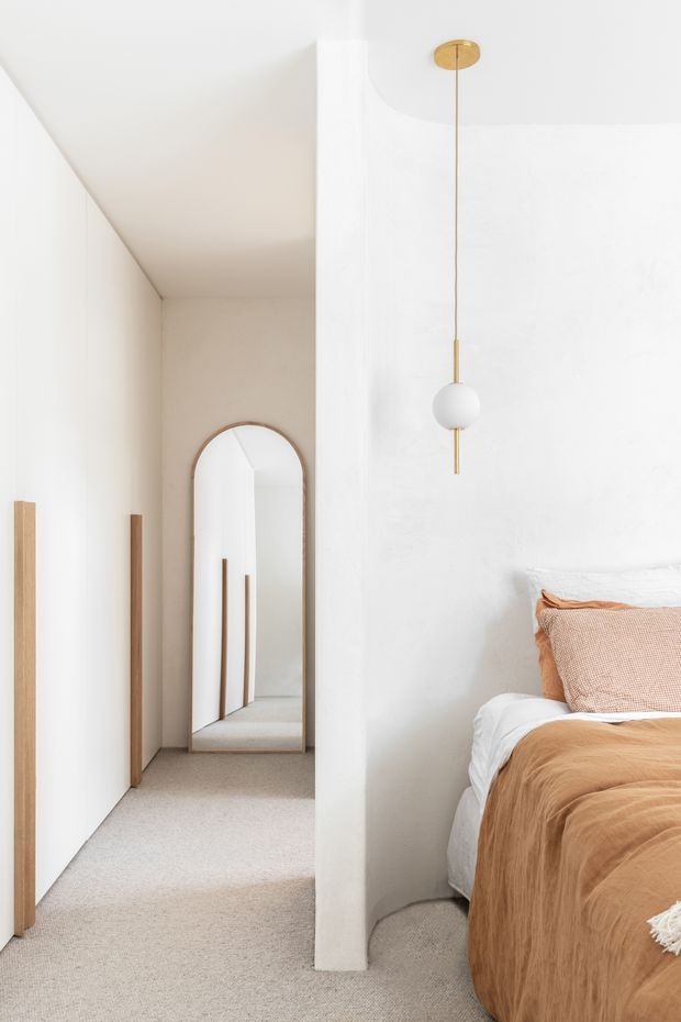 A bright, minimalist bedroom viewed toward a narrow hallway. The room has white plaster walls, a soft beige carpet, a partially visible bed dressed in warm camel-brown bedding, an arched full-length mirror, slim timber door or wardrobe panels, and a simple white globe pendant light with a brass-toned ceiling mount. The space feels calm and refined, with clean architectural lines and restrained natural tones.