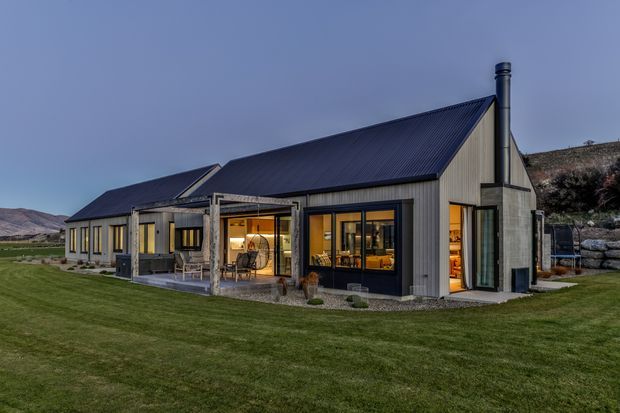 A modern single-storey rural home photographed at dusk, featuring a dark pitched metal roof, light vertical cladding, extensive black-framed glazing, and a covered outdoor terrace. Warm interior lighting contrasts with the cool blue exterior light. The house sits in a broad manicured lawn with rocky hills visible in the background.