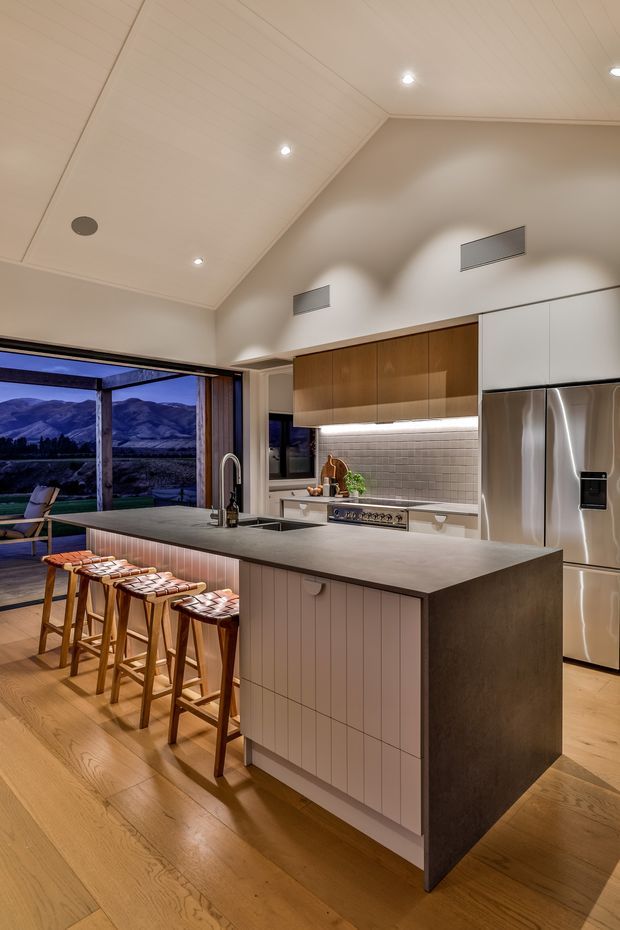 A warm, contemporary kitchen with a large central island, integrated sink and curved faucet, seating for four on timber stools, timber flooring, white vertical-panel island cabinetry, stone-look benchtops, timber overhead cabinetry, tiled splashback, and stainless-steel appliances. The kitchen opens to a glazed outdoor-facing living area with mountain views. A vaulted, white-lined ceiling with recessed lighting adds height and spaciousness.