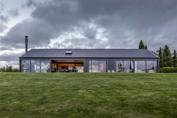 A low, single-storey contemporary house set behind a broad lawn. The dark corrugated metal gable roof and matching dark vertical-clad facade are paired with extensive floor-to-ceiling glazing, including a recessed central opening that reveals a warm-toned interior. A chimney rises at the left, and the dramatic cloudy sky creates a subdued rural or suburban setting.