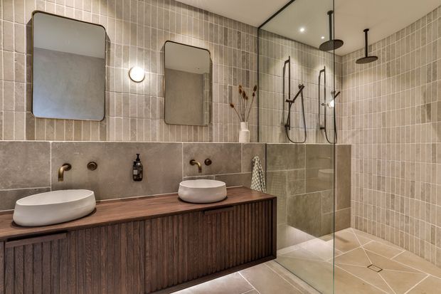 A warm, contemporary bathroom with a double vanity featuring round vessel basins on a dark fluted timber cabinet. Beige and taupe tiled walls and floor continue into a glass-enclosed walk-in shower with dual rain showerheads and brushed brass fixtures. Rounded rectangular mirrors, wall sconces, recessed lighting, and subtle decorative details complete the refined interior.