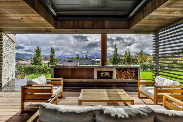 Covered outdoor entertaining area with a timber-lined ceiling, dark steel-framed roof opening, horizontal privacy screening, and a built-in outdoor kitchen or fireplace wall. Timber-framed lounge chairs and a sofa surround a low timber coffee table, while a broad lawn and mountain landscape are visible beyond the open sides. The space has a warm, contemporary lodge-like character with strong indoor-outdoor connection.