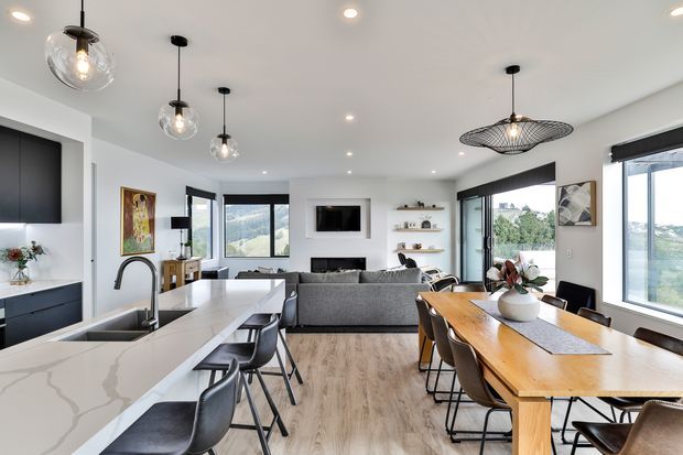 Open-plan contemporary living area combining a modern kitchen, dining space and lounge. The kitchen features a long white stone island with dark cabinetry and black bar stools, while the dining zone has a large timber table with dark chairs. A grey sofa faces a wall-mounted television, with expansive windows providing views outside. Clear glass pendant lights, recessed ceiling lighting and light timber flooring create a bright, refined interior.
