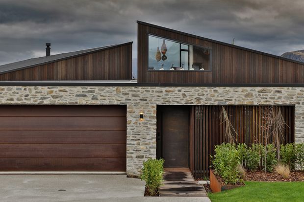 Exterior view of a contemporary single-storey residence with a sharply pitched asymmetrical roofline, dark vertical timber cladding, and a prominent natural stone masonry wall. The facade includes a wide timber-toned garage door, recessed entry, vertical timber screening, large glazing, and restrained landscaped planting under a dramatic overcast sky.