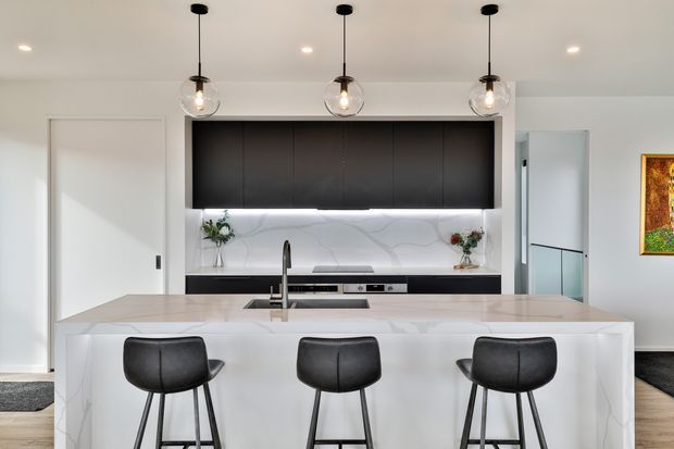 Contemporary kitchen interior dominated by a large white stone island with integrated sink and three black bar stools. The kitchen features matte black overhead cabinetry, a white veined stone splashback and benchtop, black tapware and appliances, and three globe pendant lights. White walls and ceiling create a bright, clean, high-contrast space, with a hallway or adjoining room visible beyond.