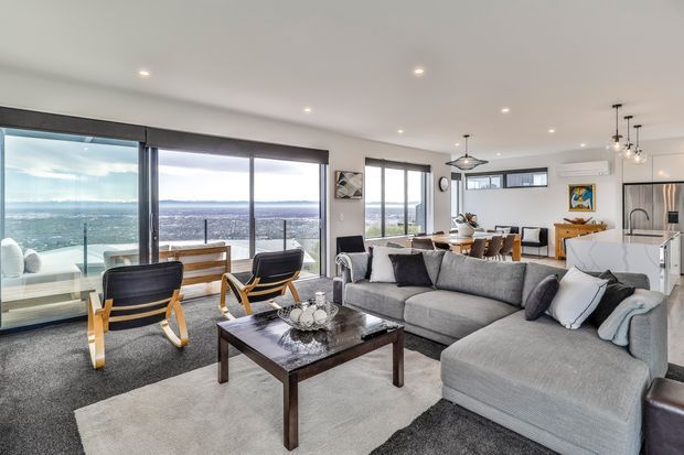 Bright, open-plan contemporary living area with a large grey sectional sofa, dark wood coffee table, lounge chairs, and a dining area beyond. Floor-to-ceiling glazing opens to expansive views, while a compact kitchen with an island is visible at the rear. Recessed ceiling lights and pendant fixtures create a refined, airy interior.