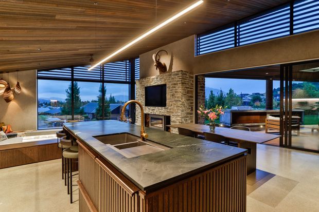 A spacious, open-plan contemporary kitchen with a large central island featuring a dark stone benchtop, integrated sink, and timber slatted cabinetry. The room has a warm timber-lined ceiling, exposed stone feature wall, expansive windows with horizontal louvres, pendant lighting, and a dining area opening to an outdoor terrace. A fireplace and television are set into the stone wall, with landscaped views beyond.