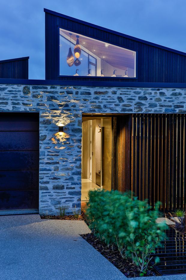 Blue-hour exterior view of a contemporary two-storey house. The facade combines dark vertical cladding, irregular natural stone masonry, a large illuminated upper-level window, a dark garage door, and a recessed timber-lined entry beside a vertical slatted screen. The driveway, landscaped planting, and warm interior lighting create a dramatic evening presentation.