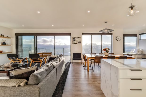 Open-plan contemporary interior combining a living room, dining area and kitchen. The room features floor-to-ceiling glazing with expansive water and city views, a large grey sectional sofa, timber dining furniture, a white island with subtle veining, recessed ceiling lights, and sculptural pendant fixtures. The palette is light and neutral with warm timber flooring and refined modern furnishings.