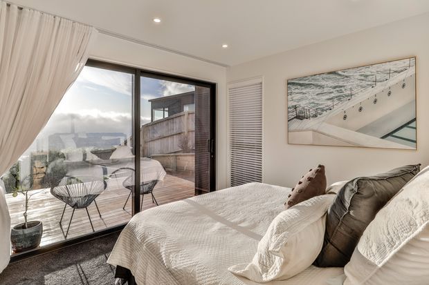 Bright, airy bedroom with a large bed dressed in white textured bedding, neutral pillows, sheer white curtains, dark carpet, and expansive sliding glass doors opening to a timber deck. A coastal-looking architectural artwork hangs on the wall, while the glazing provides views toward neighbouring structures and distant landscape.