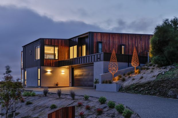 A contemporary multi-level home set into a sloping landscaped site, photographed at dusk. The house features dark metal cladding, warm vertical timber-look wall sections, extensive glazing, a projecting upper volume, an integrated garage, and a dark metal balcony or terrace balustrade. Low planting and sculptural illuminated outdoor forms line the driveway and retaining walls.