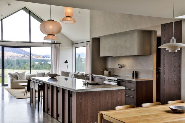 Open-plan contemporary kitchen and dining area with a large dark timber island, pale stone benchtops, integrated cabinetry, stainless-steel appliances, and a warm timber dining table. Floor-to-ceiling glazing and a high angled ceiling provide expansive mountain views and abundant natural light. Pendant lighting and minimal furnishings create a refined, warm modern interior.