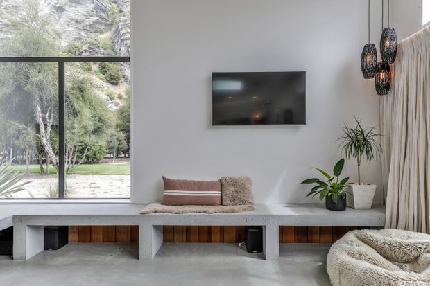 Bright contemporary living room with a wall-mounted television, a long built-in concrete bench featuring warm timber insets, indoor plants, woven pendant lights, and full-height glazing overlooking greenery. Neutral finishes and textured furnishings create a calm, natural atmosphere.