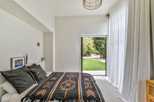 Bright, calm bedroom with a neatly made bed, dark patterned throw and textured cushions, white walls, sheer floor-to-ceiling curtains, a woven pendant light, and a full-height glazed door opening to a green garden.