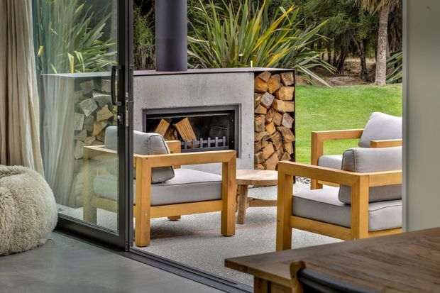 View from an interior living area through fully opened floor-to-ceiling glazing to a covered outdoor seating area. The outdoor space features a built-in concrete fireplace with stacked firewood, timber-framed upholstered armchairs, a small timber side table, and lush garden planting beyond. A dark timber dining table is partially visible inside, with a textured rug and neutral furnishings.