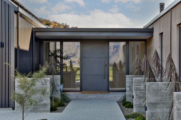 A contemporary single-storey entrance courtyard framed by two dark metal-clad wings. The central recessed entry has a broad flat roof, a dark solid pivot door, and full-height glazed sidelights. Concrete or rendered rectangular planters, gravel paving, and sculptural, drought-tolerant landscaping create a restrained modern approach.