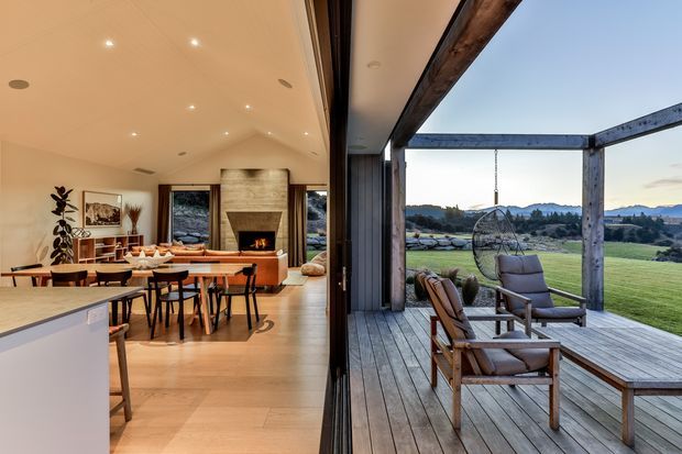 A warm, contemporary open-plan living and dining area viewed alongside a spacious timber deck. The interior features pale timber flooring, a long dining table, kitchen island, recessed ceiling lights, large glazing, and a stone-clad fireplace with a firebox. The deck has timber decking and exposed timber posts and beams, with suspended outdoor seating overlooking a rural landscape.