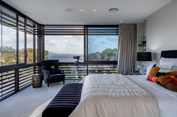 Contemporary bedroom with a large upholstered bed, black channel-tufted bench, armchair, and floor-to-ceiling corner glazing overlooking a coastal landscape. Dark window frames, layered curtains, recessed ceiling lights, and a telescope create a refined retreat with abundant natural light.