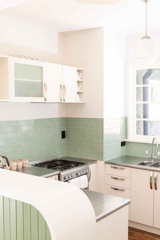 Bright compact kitchen with white cabinetry, brushed metal handles, pale mint-green subway tile splashback, stainless-steel benchtops and appliances, and a curved peninsula with vertical green panelling. A white-framed window and pendant light add natural light to the space.