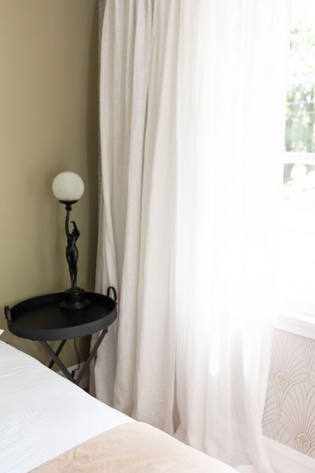 A softly lit bedroom corner featuring a neatly made bed in the foreground, a small dark round side table with a sculptural lamp, olive-beige painted wall, and full-height sheer cream curtains beside a bright window. Decorative patterned radiator or wall panel detail is visible below the window.