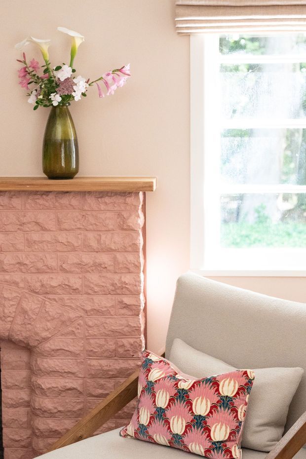 A softly lit residential interior corner featuring a textured painted brick fireplace with a timber mantel, a tall green vase of flowers, a white-framed window with a woven Roman blind, and a light upholstered armchair with a colourful floral cushion. The palette is warm, pastel, and inviting.