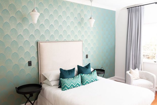 Bright, stylish bedroom featuring a large upholstered cream headboard with nailhead trim, white bedding, and turquoise patterned accent cushions. The feature wall has an aqua and teal scallop-patterned wallpaper, complemented by matching pendant lights, dark bedside tables, and full-length grey curtains beside a bright window. A white upholstered armchair sits in the corner.