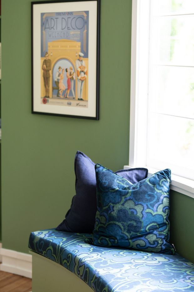 A compact window seat or bench upholstered in a bold blue, teal and green patterned fabric, with coordinating cushions. The seat is positioned beside a bright white-trimmed window against a deep sage-green wall, with a framed Art Deco poster above and timber flooring visible below.