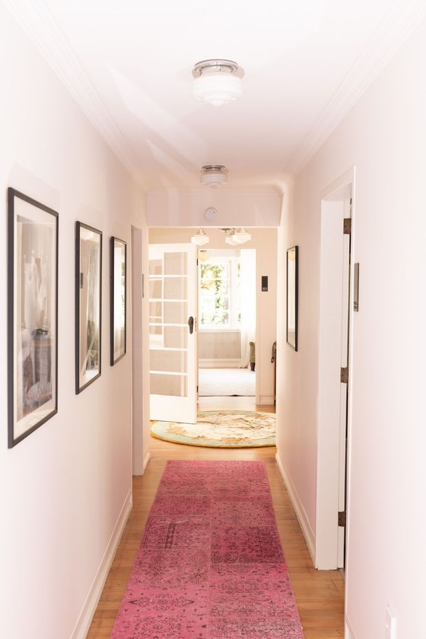 A long, narrow residential hallway with pale pink-white walls, white trim, wood flooring, framed artwork, ceiling-mounted flush lights, and colorful Persian-style runners. Open doorways lead toward a bright bedroom or sitting area at the end of the corridor.