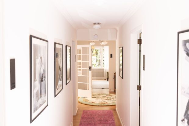 Bright, narrow residential hallway with white walls and ceiling, framed artwork, multiple doorways, and layered rugs. A partially open glazed door leads toward a bedroom or sitting area at the end of the corridor, with daylight and greenery visible beyond.