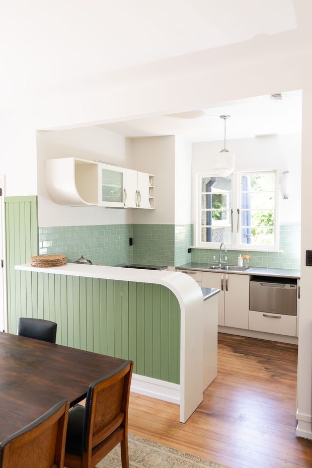 Bright, compact kitchen viewed from an adjoining dining area. The kitchen features pale cabinetry, a curved white breakfast-bar surround with vertical sage-green panelling, a glossy mint-green subway-tile splashback, stainless-steel appliances and fixtures, timber flooring, and a large multi-pane window. A dark timber dining table and chairs occupy the foreground.