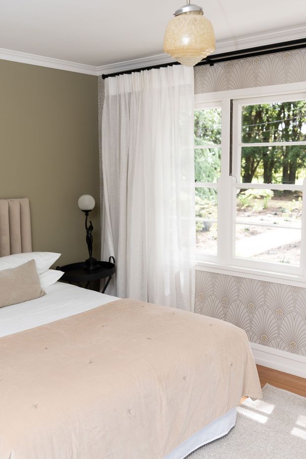 Bright, elegant bedroom with a partially visible upholstered bed dressed in white bedding and a soft beige coverlet. The room features muted taupe painted walls, white crown moulding, sheer white curtains, a black bedside table and lamp, a textured pendant light, and distinctive Art Deco-inspired fan-pattern wallpaper around the window. White-trimmed glazing overlooks leafy landscaping, while pale timber flooring and a light rug complete the calm, refined scheme.