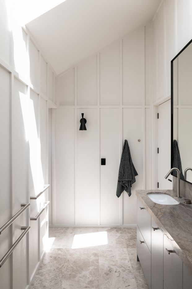 Bright, narrow contemporary bathroom with white vertical wall panelling, a pale stone tiled floor, and a long white vanity topped with a grey-veined stone benchtop. A wall-mounted black sconce, black towel, brushed metal tapware, and a large frameless mirror add contrast. Strong sunlight enters from the left, creating an airy, refined atmosphere.