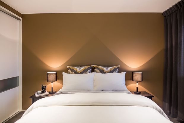 A cozy, symmetrically arranged bedroom with a neatly made bed, white bedding, patterned accent cushions, dark bedside tables, matching table lamps, and a warm brown feature wall. Soft uplighting creates geometric triangular shadows behind the bed, while dark full-length curtains frame the right side and a built-in wardrobe is visible on the left.