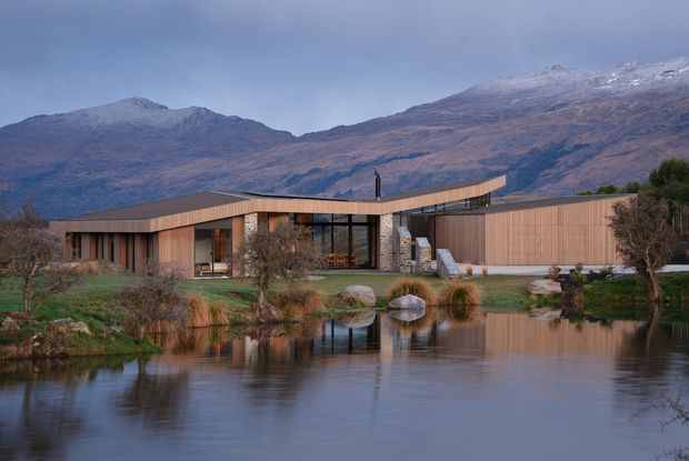 A low, contemporary timber-clad residence set beside a reflective pond, with broad glazed openings, a gently folded roofline, landscaped grounds, and rugged snow-dusted mountains in the background. The building has a warm natural palette and appears designed to frame the surrounding alpine landscape.