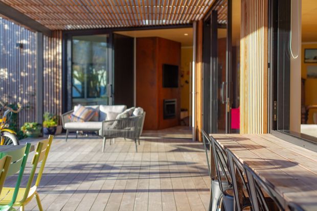 A sunlit contemporary indoor-outdoor living and dining area with large glazed openings, timber slatted screening and a pergola-style ceiling. The tiled terrace contains a lounge setting and a long timber dining table with chairs, while a warm timber-lined living room and media wall are visible beyond.