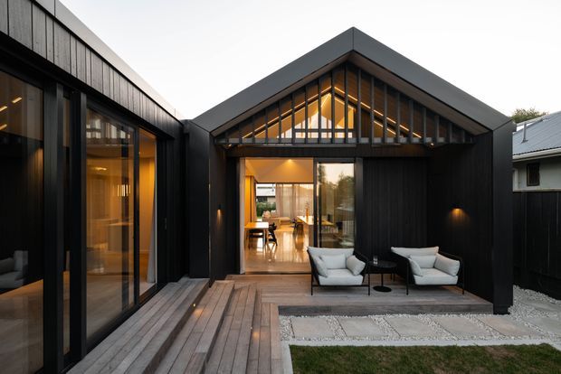 Contemporary dark-clad residential courtyard viewed from an outdoor timber deck. The central gabled pavilion has a glazed triangular clerestory and large sliding glass doors opening into a warmly lit dining and kitchen area, while a connected wing sits to the left. Two outdoor lounge chairs occupy the deck, with gravel paving and lawn in the foreground.