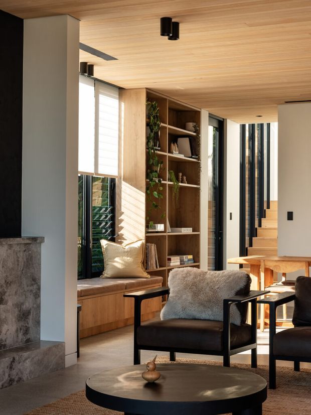 A warm contemporary living room with a timber-lined ceiling and built-in timber shelving surrounding a window seat. The space includes dark leather lounge chairs, a round black coffee table, a timber dining table in the background, large glazed openings, and a staircase leading upward. Natural light highlights the restrained, refined material palette and indoor planting.