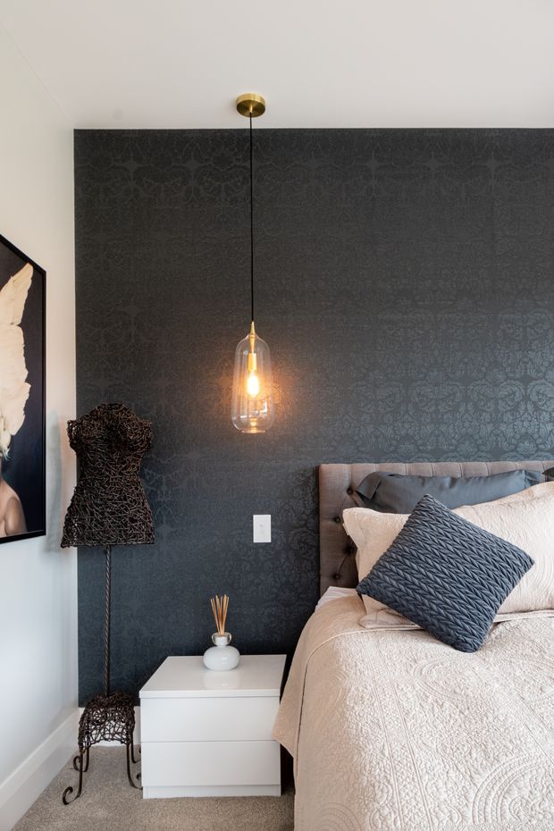 A stylish bedroom corner featuring a tufted grey upholstered bed, layered cream bedding, and a dark patterned feature wall. A warm pendant light hangs centrally above a white bedside table, while a sculptural wire mannequin and framed artwork add decorative accents.