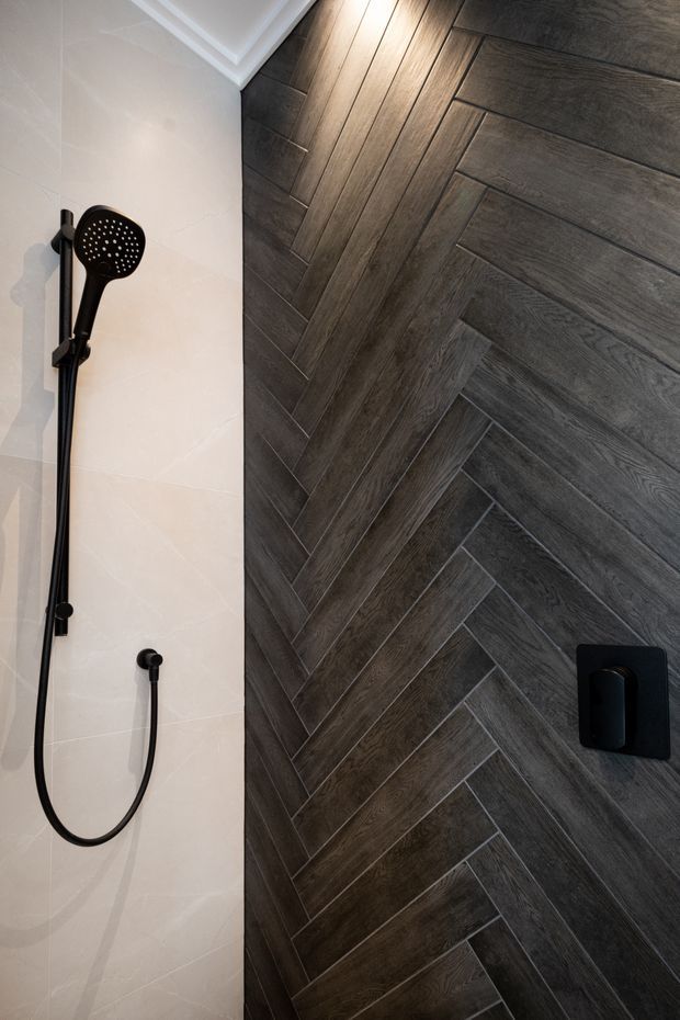 Close-up view of a contemporary shower enclosure featuring a black handheld shower fixture and controls mounted on a pale tiled wall, alongside a dramatic dark wood-look herringbone tile feature wall. White ceiling trim is visible above.