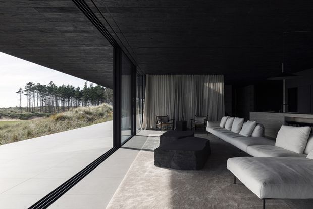 A dramatic, dark-toned contemporary living room opening onto a broad outdoor terrace and grassy coastal-looking landscape. Full-height sliding glass doors, a black ceiling, dark walls and cabinetry, pale sectional seating, a large dark ottoman, and floor-to-ceiling curtains create a minimalist, high-contrast interior with strong indoor-outdoor connection.