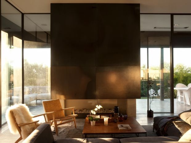 A warm, refined living room with floor-to-ceiling glazing and dark-framed glass partitions. A large dark metal-clad fireplace or feature wall dominates the centre, with a low timber coffee table, mid-century-style timber lounge chairs, upholstered seating, and a dining area visible beyond. Natural light, garden views, and earthy materials create a calm contemporary atmosphere.