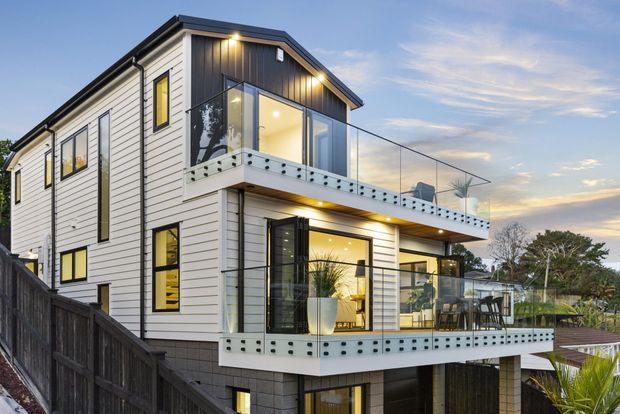 Contemporary multi-level residential house viewed from outdoors at dusk, featuring white horizontal cladding, dark trim, extensive glazing, two projecting balconies with clear glass balustrades and perforated solid panels, and warm interior lighting. The home sits on a sloping suburban site with landscaped surroundings.