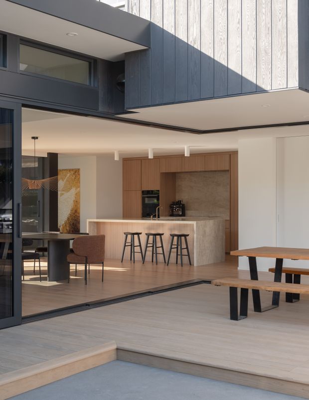 A contemporary indoor-outdoor entertaining area viewed from a sunken exterior terrace, with a large open kitchen and dining space beyond full-height dark-framed glazing. The kitchen features warm timber cabinetry, a stone island with three stools, integrated appliances and a neutral stone splashback. A timber dining table and bench sit on a light timber deck, while dark vertical exterior cladding and strong cantilevered roof forms frame the space.
