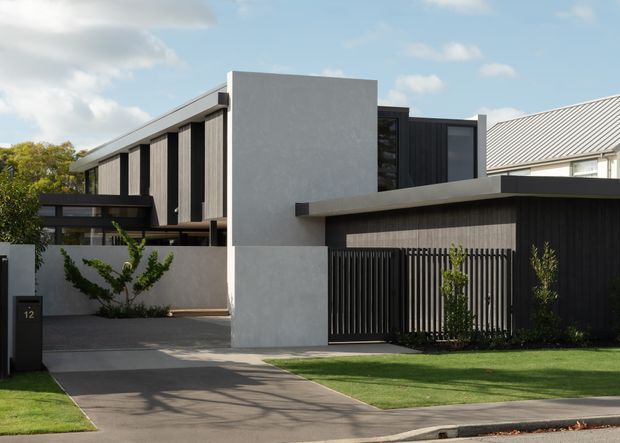 Street-facing view of a contemporary multi-level residential house with strong geometric massing, a flat-roofed dark garage volume, light rendered concrete-like walls, vertical black screening, and large dark-framed glazing. The property includes a driveway, lawn, front garden, and a contrasting pitched metal roof visible behind the main structure.