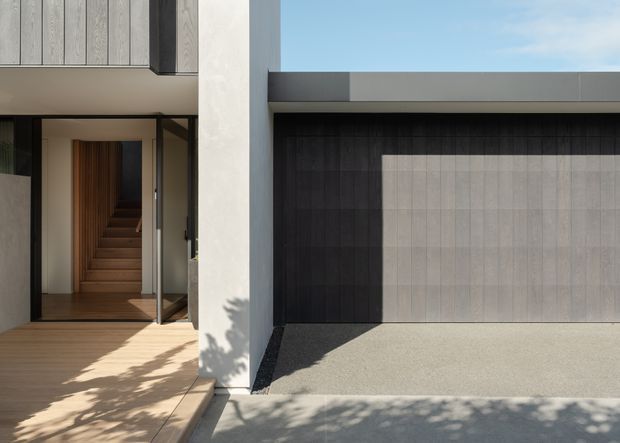 Contemporary residential facade viewed from outside, featuring a recessed entry with timber steps and panelling, pale rendered walls, dark vertical garage cladding, and a broad light grey garage door. Strong sunlight creates crisp shadows across the timber porch and concrete driveway.