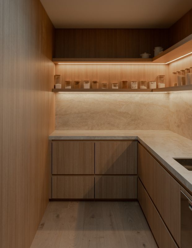 Compact, warm-toned kitchen or pantry with continuous timber cabinetry, a pale stone benchtop and matching backsplash, integrated under-shelf lighting, open display shelving with jars, and timber flooring. The L-shaped joinery and partially visible sink indicate a highly resolved small kitchen fit-out.